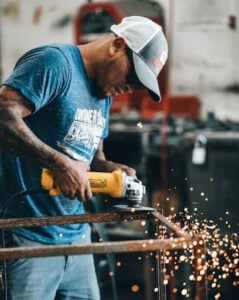A welder wearing a blue T-shirt, jeans, and a white baseball cap uses a handheld grinder to cut metal, producing bright sparks.