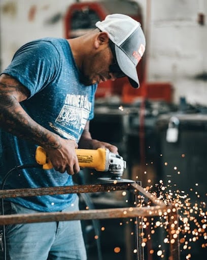 A welder wearing a blue T-shirt, jeans, and a white baseball cap uses a handheld grinder to cut metal, producing bright sparks.