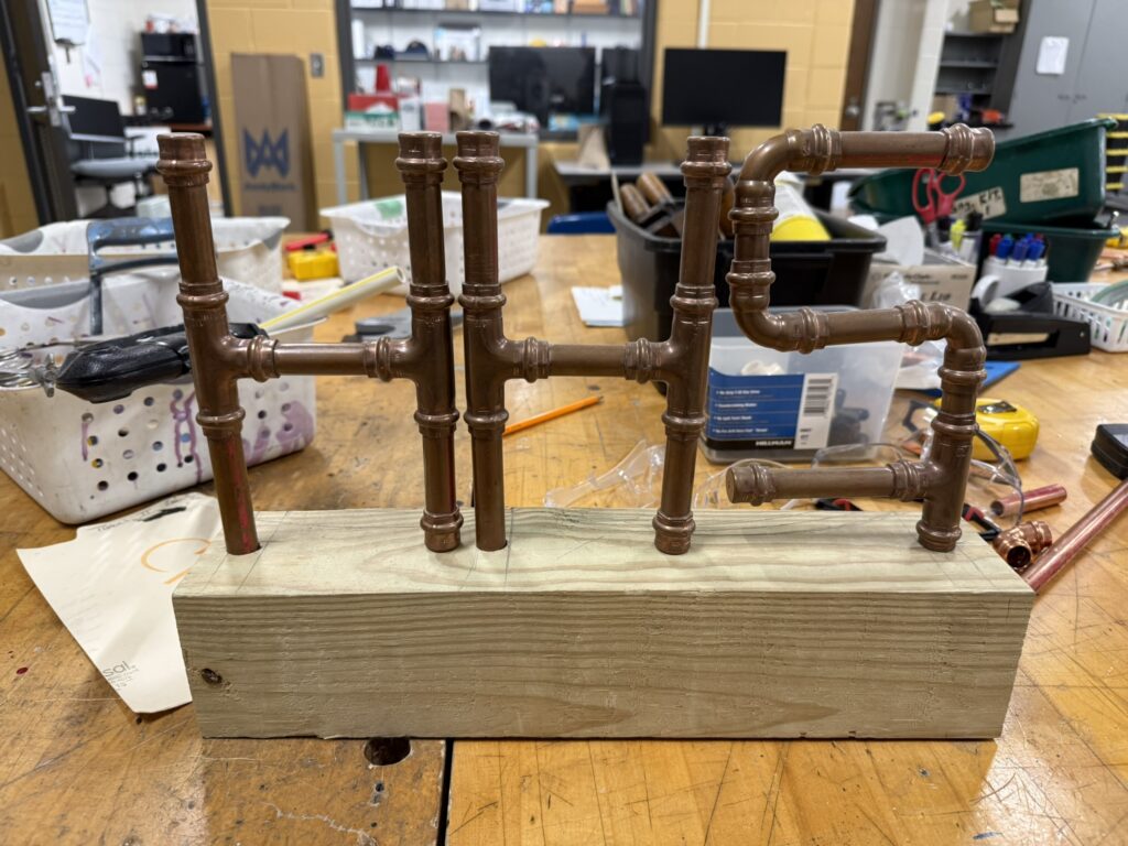 A student-built plumbing pipe assembly with copper and brown pipes mounted on a wooden base in a high school trades classroom.