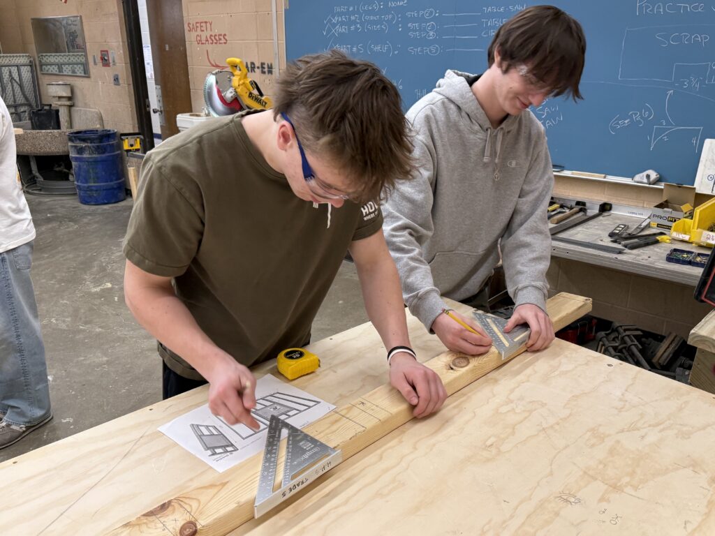 Two students wearing safety glasses measure and mark a wooden board in a trades classroom workshop.