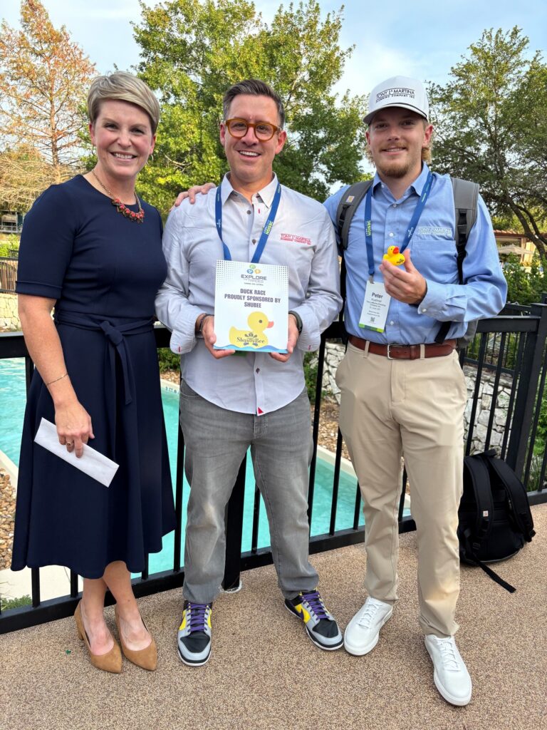 Three people smile together at an outdoor Duck Race event, one holding a sponsor thank-you sign and another holding a yellow rubber duck.