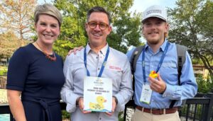 Three people smile together at an outdoor Duck Race event, one holding a sponsor thank-you sign and another holding a yellow rubber duck.