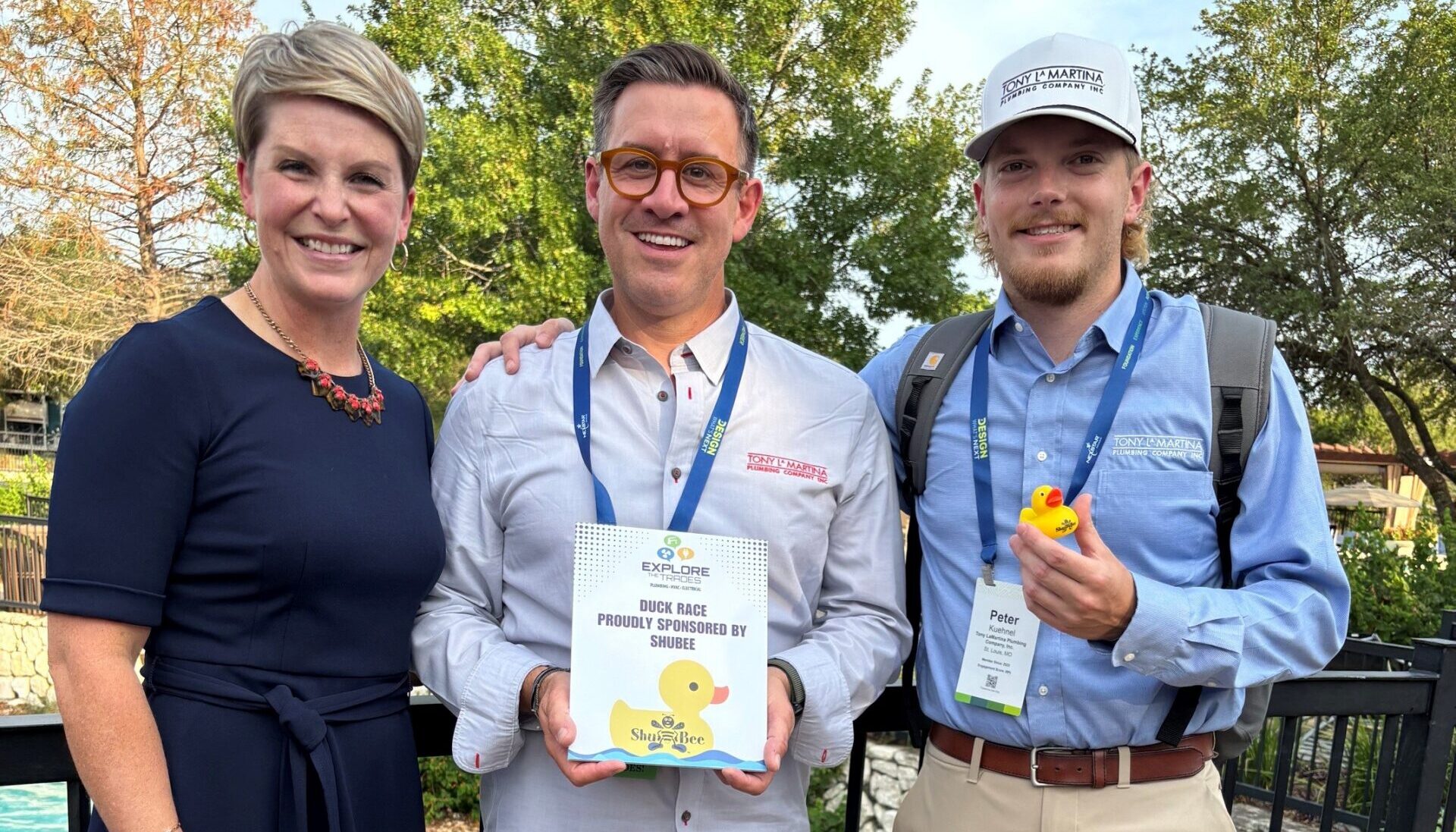 Three people smile together at an outdoor Duck Race event, one holding a sponsor thank-you sign and another holding a yellow rubber duck.