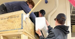 Three students work together in a shop classroom to mount a ductless mini-split indoor unit onto a wood-framed training wall.